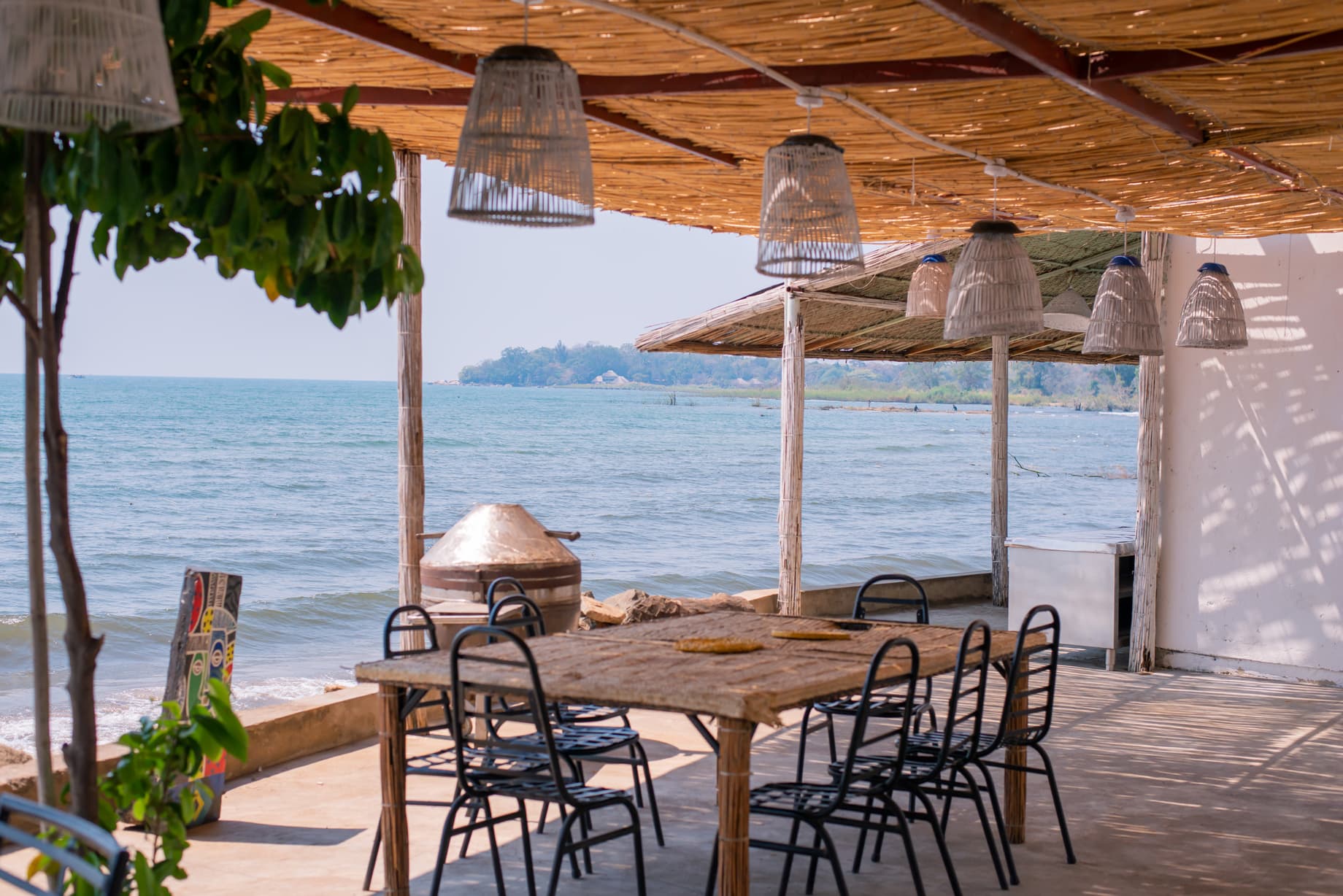 Rustic outdoor dining table with chairs overlooking Lake Malawi under a thatched roof