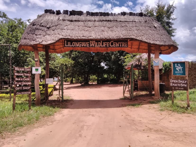 Entrance gate to a wildlife centre with thatched roof, wooden signs, and a dirt path surrounded by greenery.